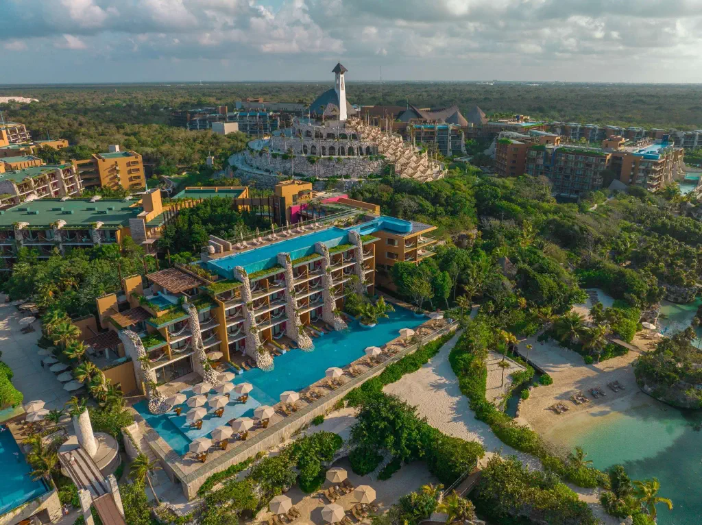 Aerial view of a luxury resort with pools, lush greenery, and modern architecture surrounded by tropical landscape and sandy beach.