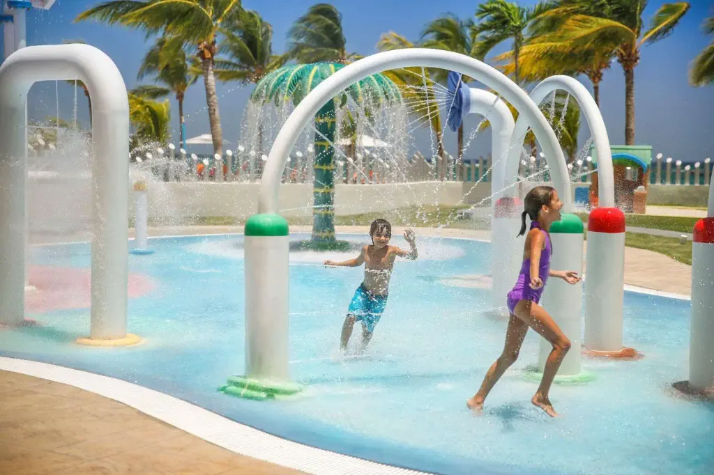 Children joyfully play in a water park, running through arched fountains. The scene is lively and fun, with palm trees and a clear sky in the background.