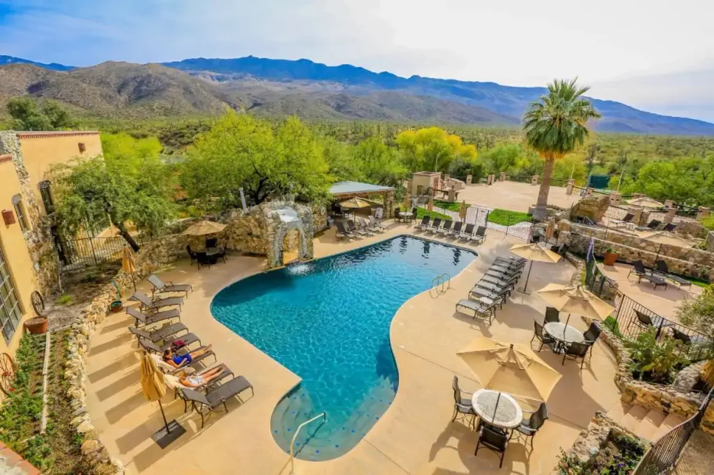 A serene resort pool with sun loungers, umbrellas, and a waterfall feature is surrounded by lush trees and mountains under a clear blue sky.