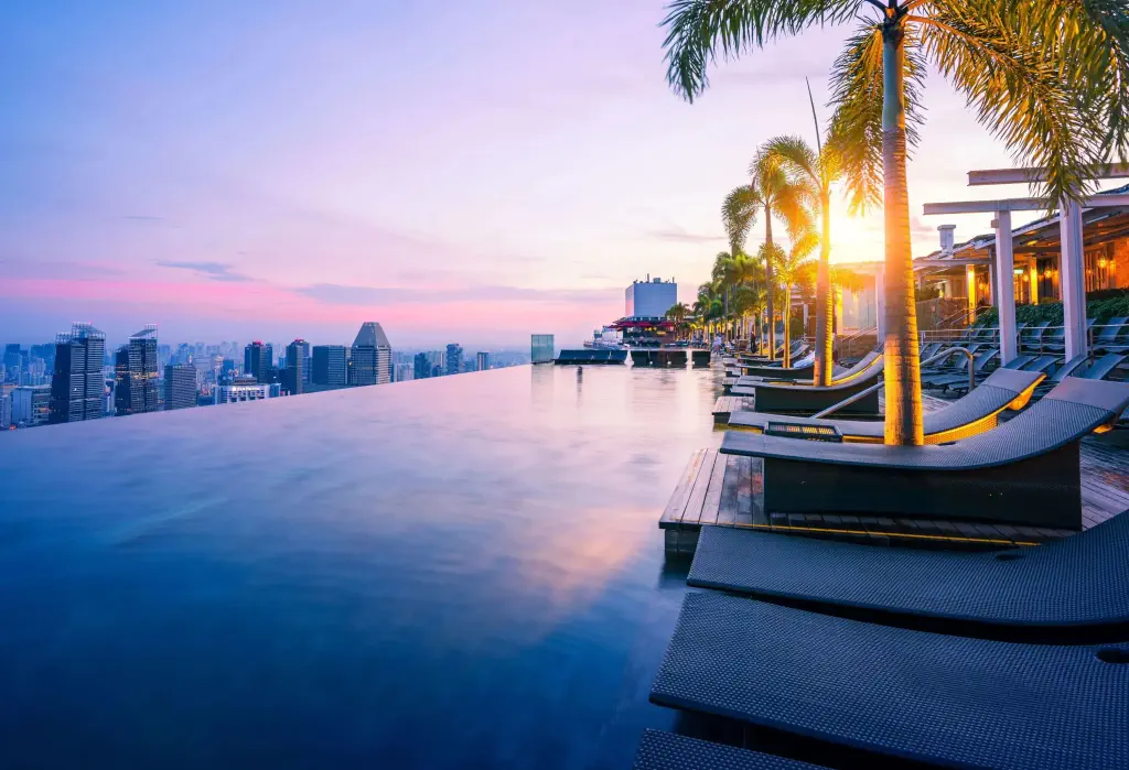 A dazzling rooftop infinity pool lined with sun loungers under the sunlit palm trees and a stunning city skyline view.