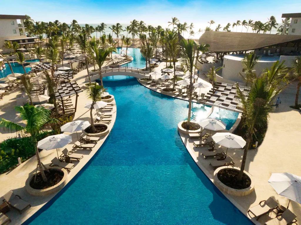 Aerial view of a luxury resort pool with lounge chairs, white umbrellas, and palm trees lining a winding blue pool. A small bridge spans the pool. Serene and inviting atmosphere.