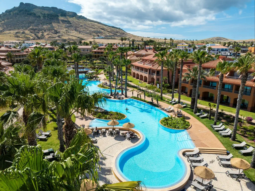 Aerial view of a resort with winding turquoise pools, surrounded by palm trees and sun loungers. Terracotta-roofed buildings are nestled amid green hills.