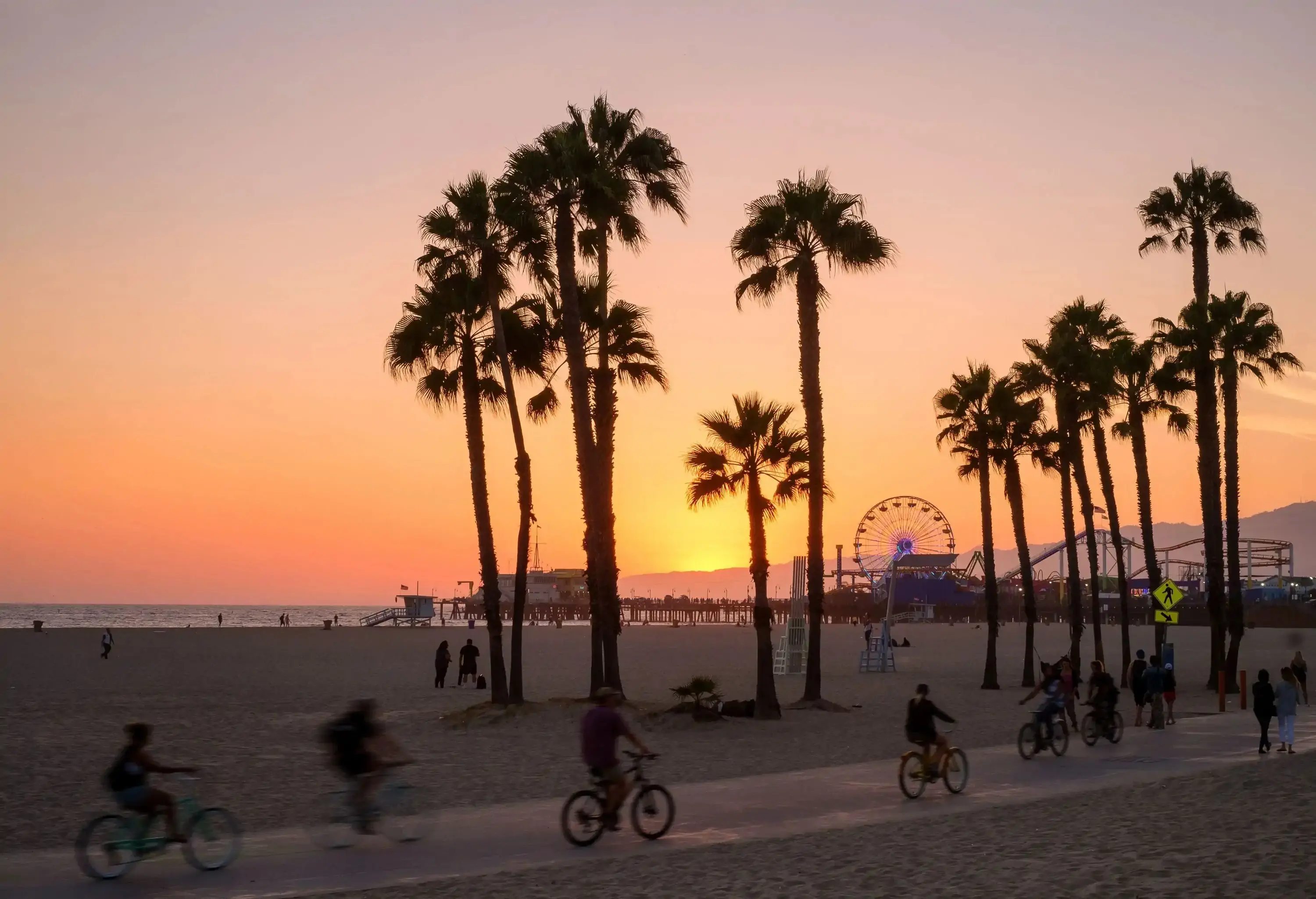 At sunset, people joyfully ride their bikes along the beachfront pathway, while a silhouette of palm trees adds a touch of serenity against the vibrant sunset backdrop, with the pier and ferris wheel visible in the enchanting background.