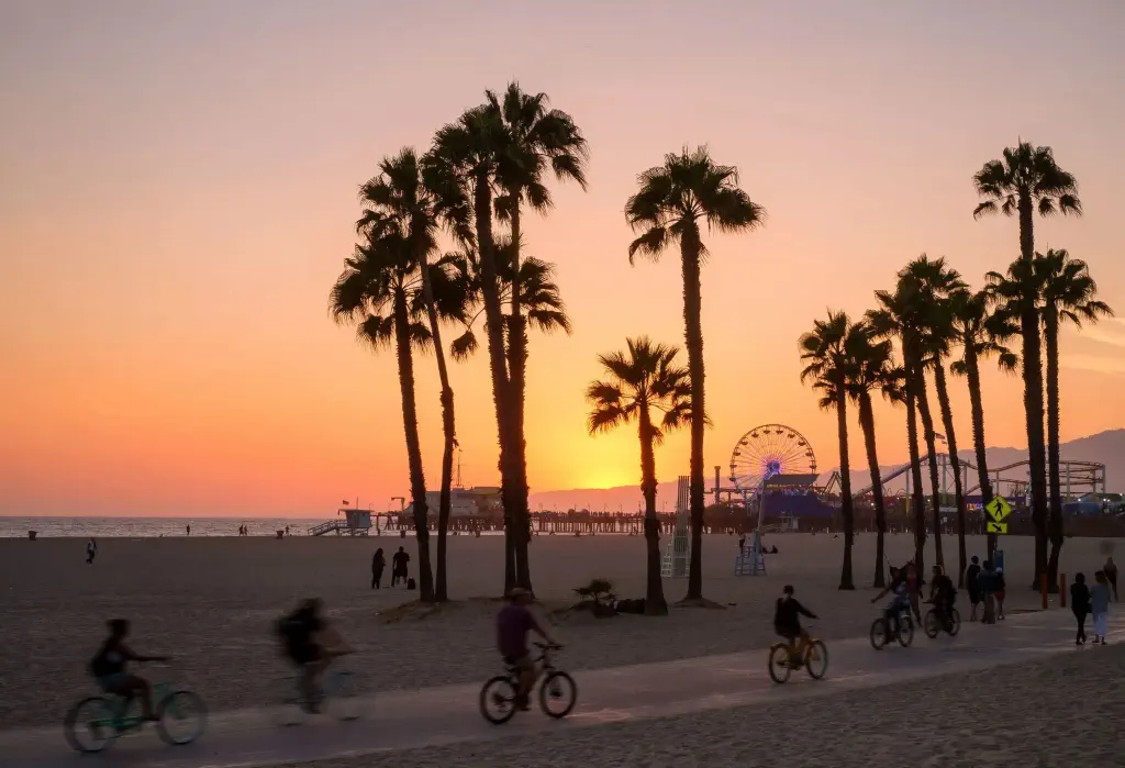 At sunset, people joyfully ride their bikes along the beachfront pathway, while a silhouette of palm trees adds a touch of serenity against the vibrant sunset backdrop, with the pier and ferris wheel visible in the enchanting background.