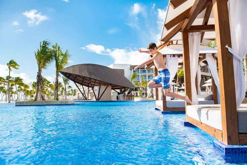 A joyful boy in blue swim trunks jumps into a bright blue pool from a wooden cabana. Palm trees sway under a sunny sky, creating a tropical vibe.