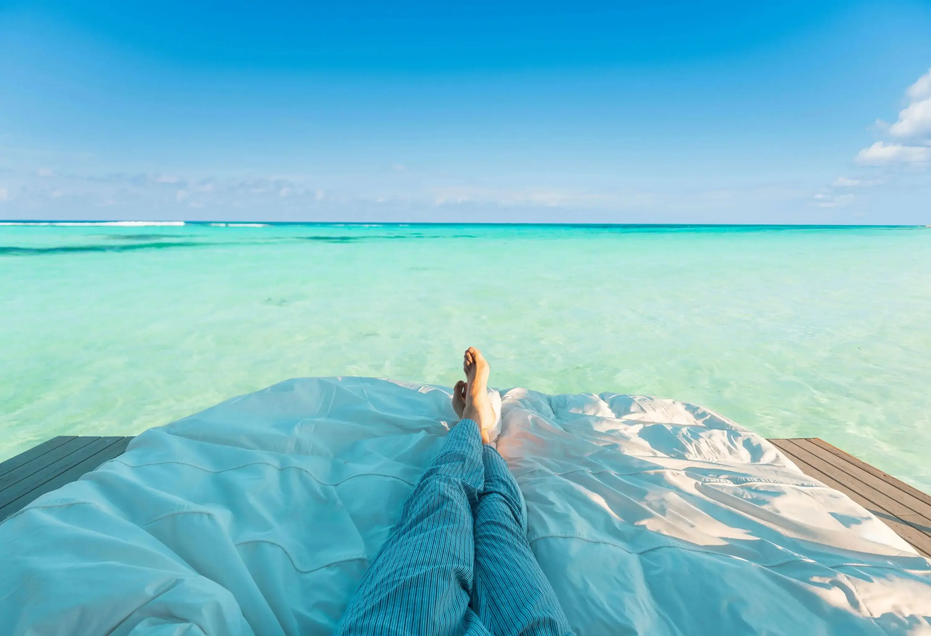 A close-up on a person's feet relaxing on an outdoor bed, with the vibrant turquoise water stretching out to the horizon beyond.