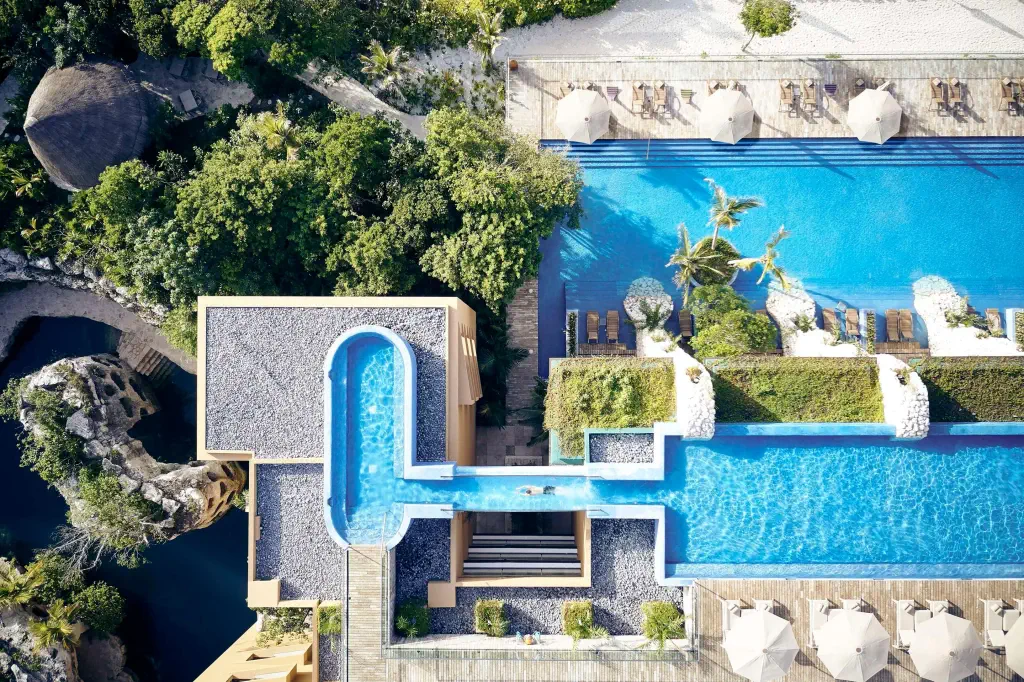 Aerial view of a luxurious resort with multiple pools, sun loungers, tropical plants, and shaded areas surrounded by greenery.