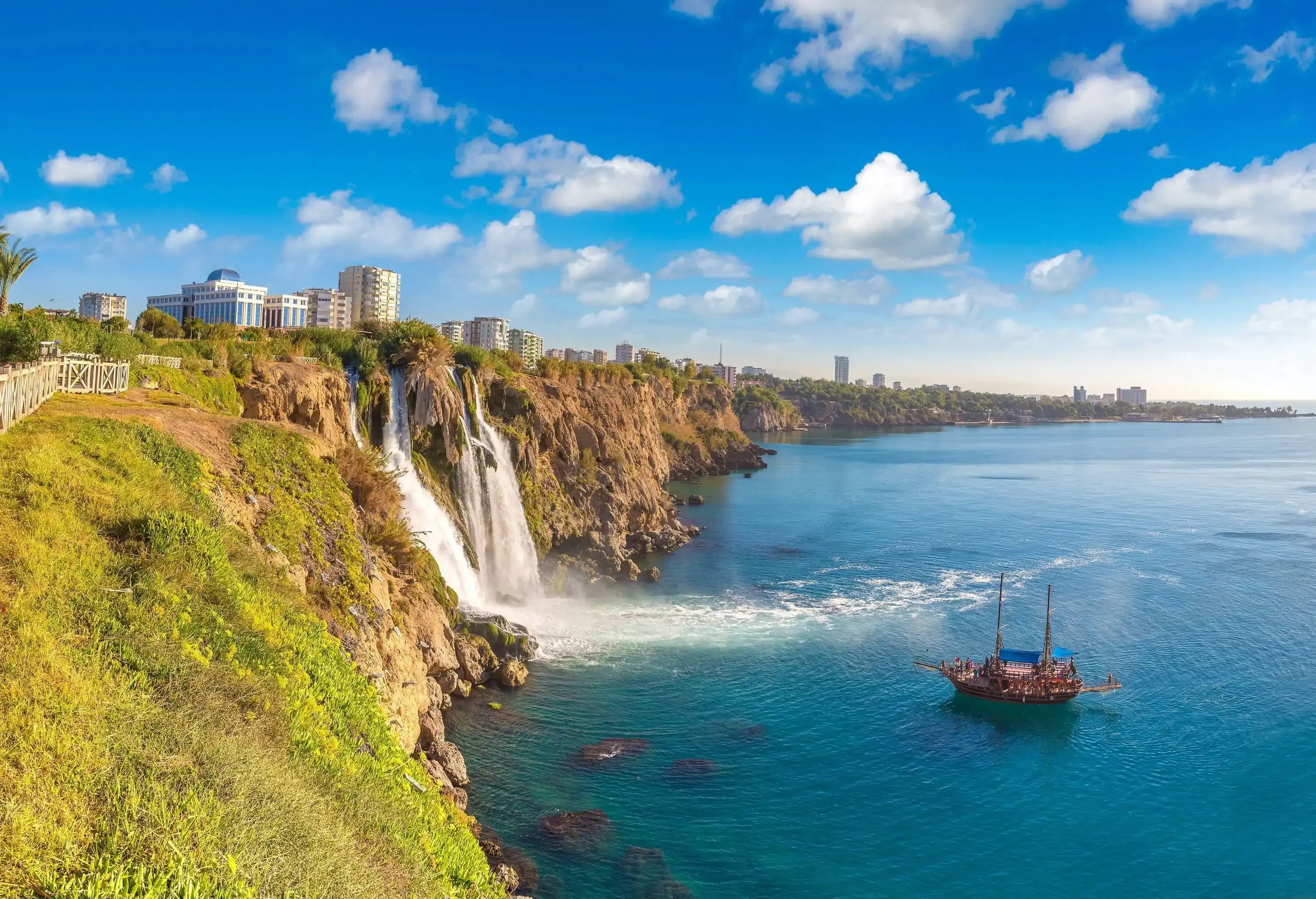A seaside waterfall pours into the ocean next to a floating boat with distant cityscape views.