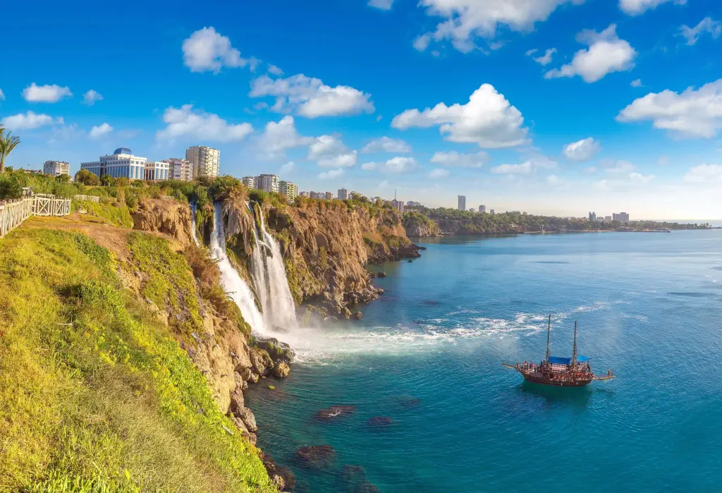 A seaside waterfall pours into the ocean next to a floating boat with distant cityscape views.