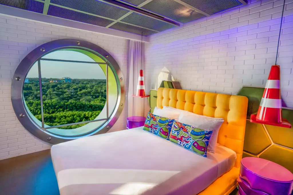 Colorful hotel room featuring a bright orange bed, Teenage Mutant Ninja Turtles pillows, and a large round window overlooking lush greenery.