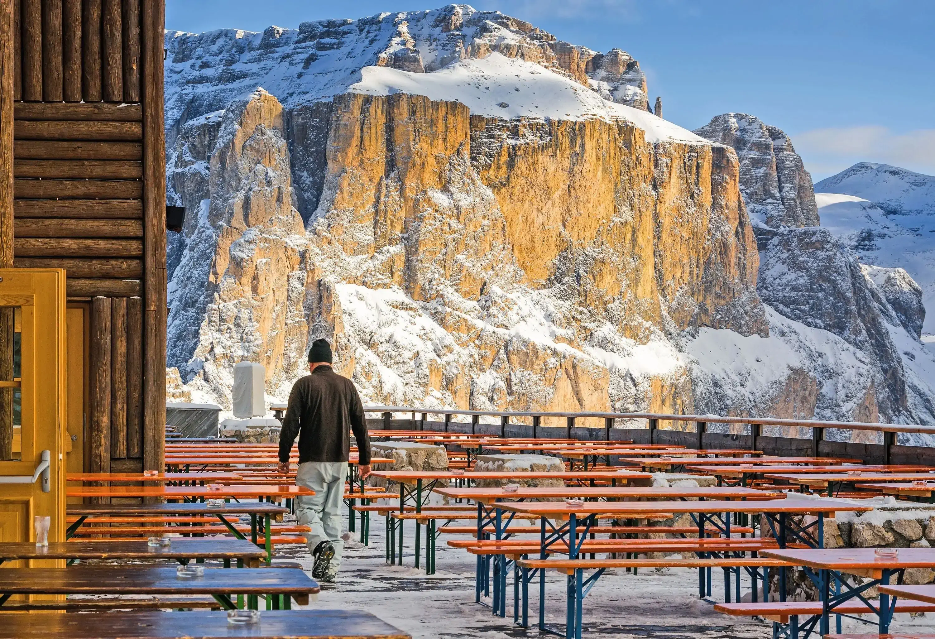 A person walks among rows of wooden chairs while gazing at a snow-capped rocky mountain.