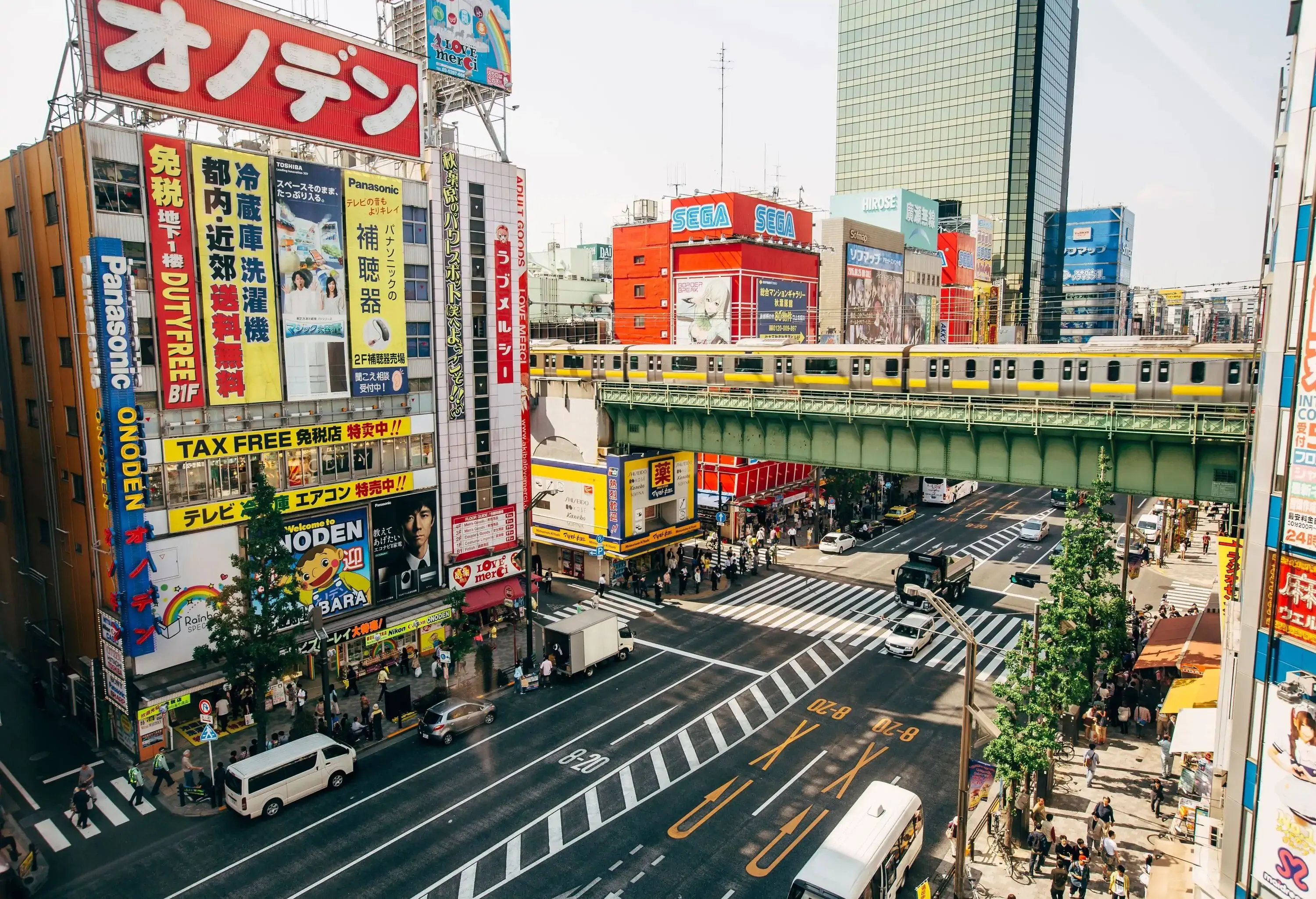 An avenue adorned with shops on both sides, featuring vibrant and colourful advertisements, while an elevated train gracefully glides along the tracks above.