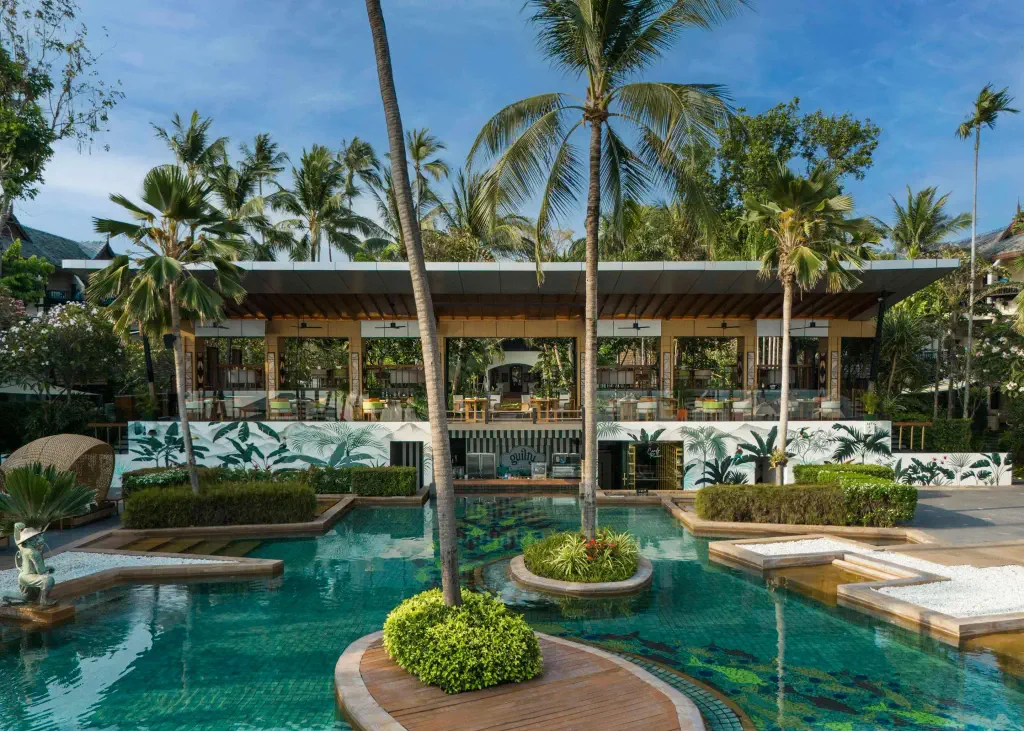 Tropical resort setting with a clear blue pool, lush palm trees, and a chic open-air building. 