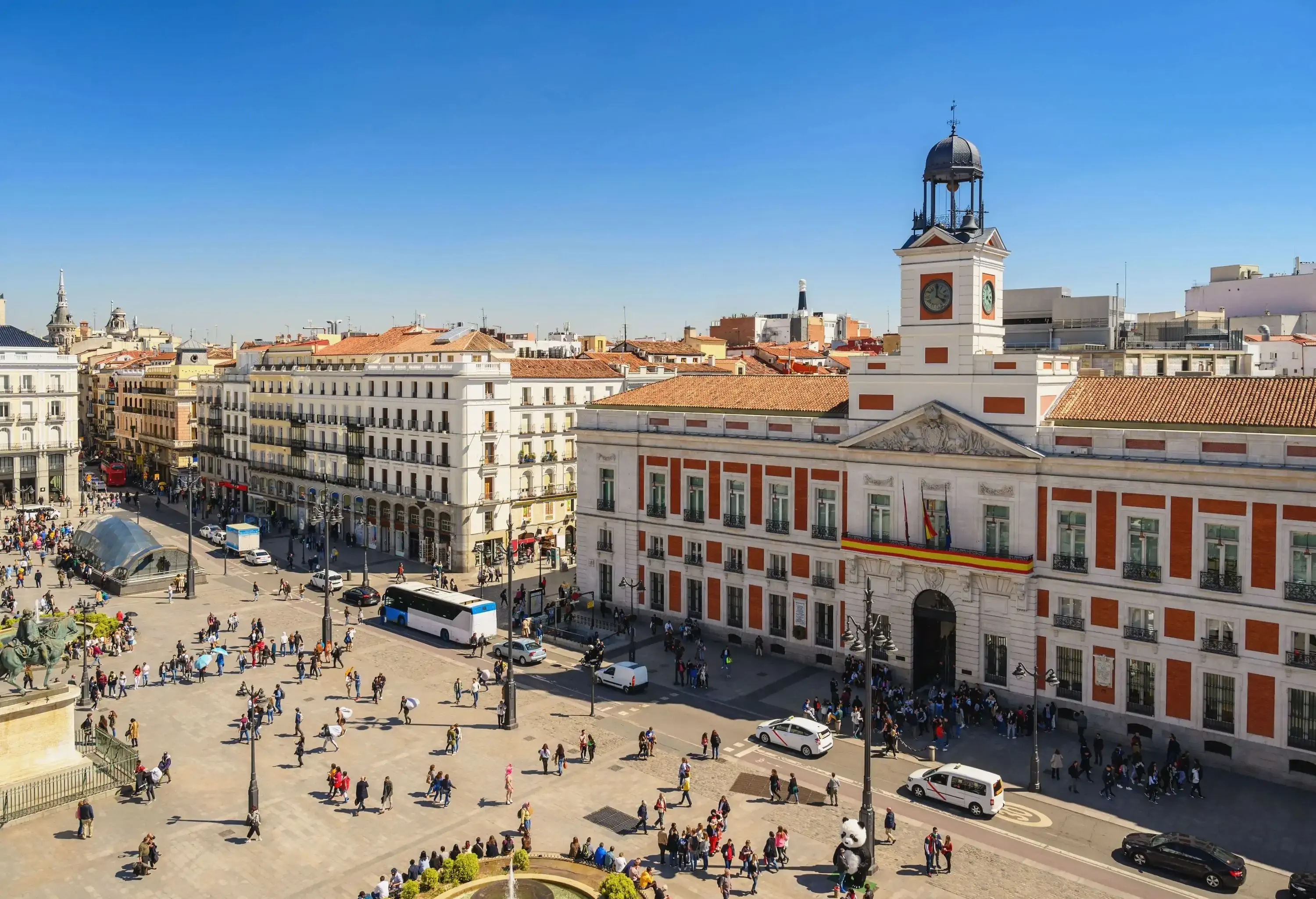 A busy public square in front of a clock tower with a bell on top.