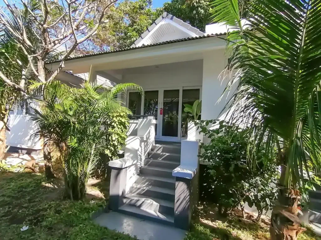Small white house surrounded by lush green palms and bushes. A shadowed staircase leads to the entrance, creating a serene, tropical feel.