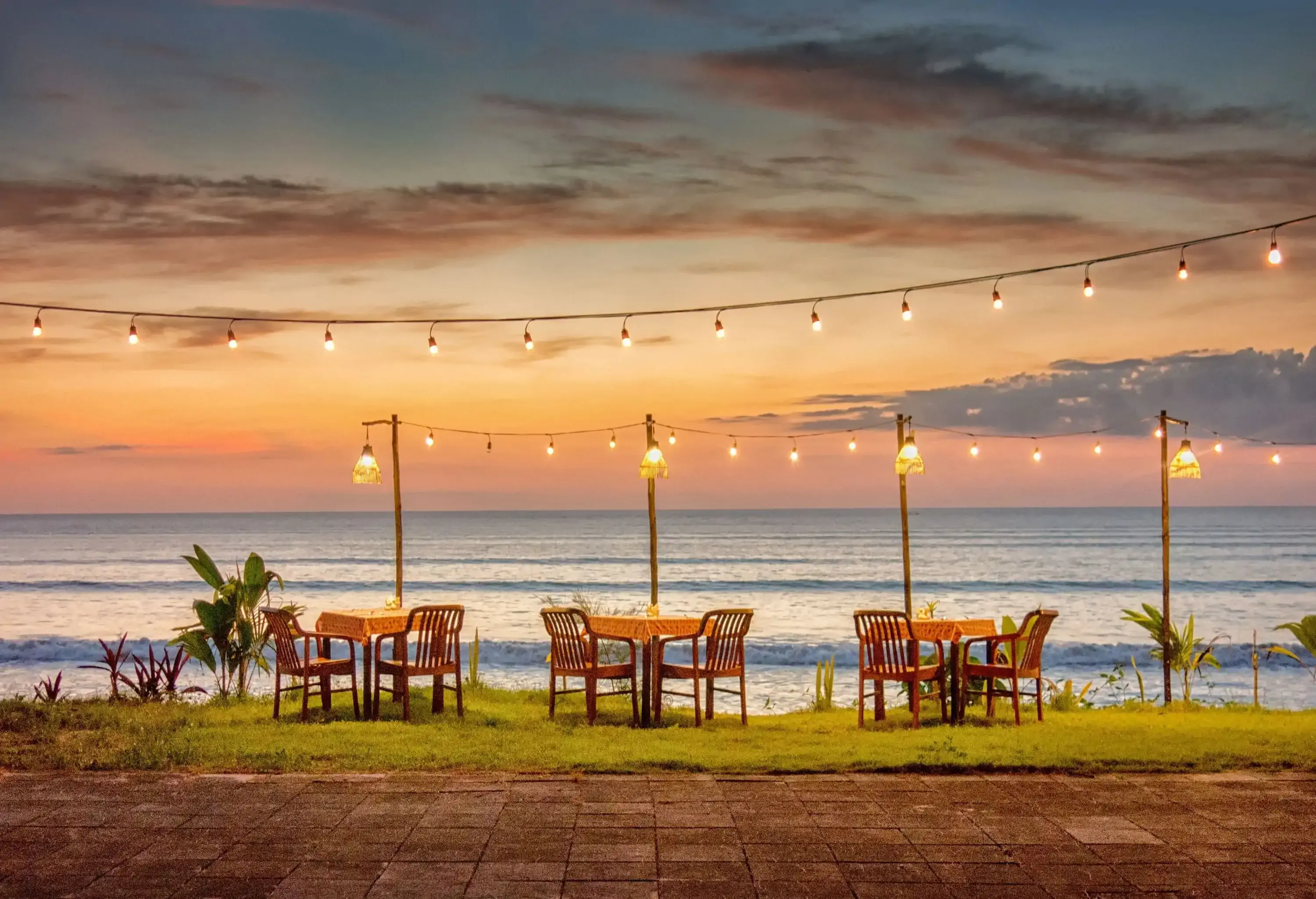 Wooden tables and chairs on the lush shore overlooking the tranquil sea against a scenic sunset sky.