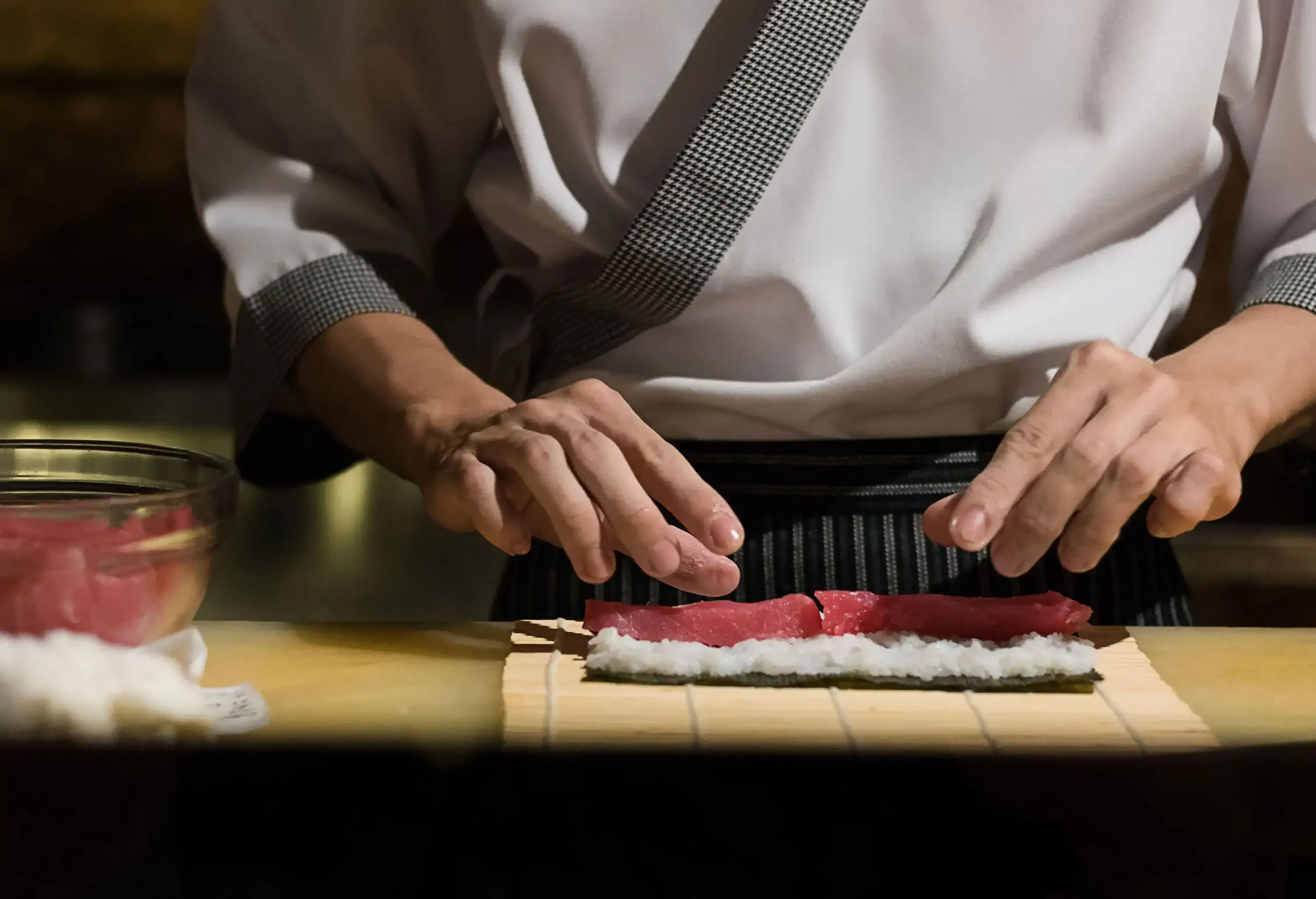 A pair of hands moulds raw fish meat, steamed rice, and seaweed into a sushi roller.