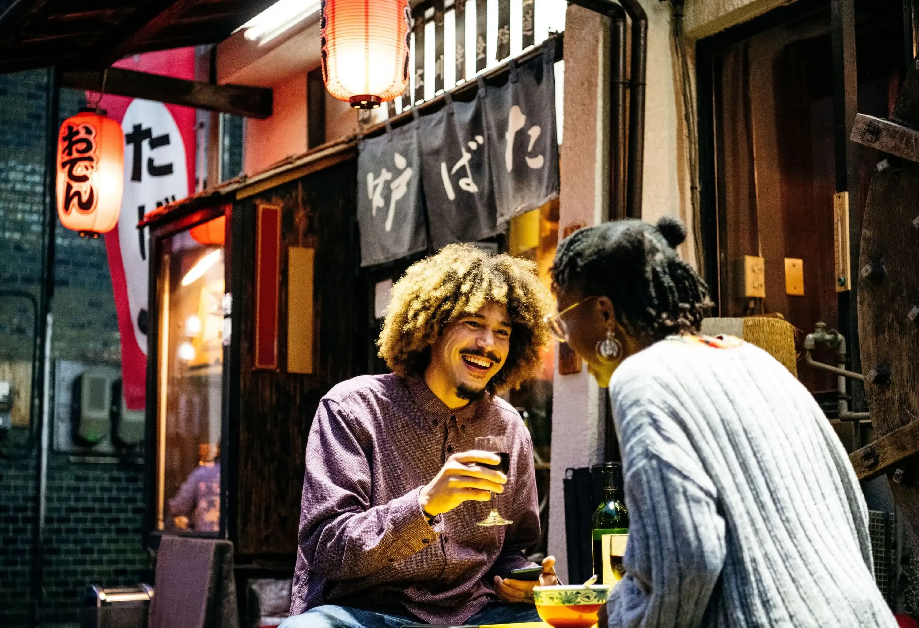 Two smiling people are seated at an outdoor table with drinks and food, in front of a building adorned with hanging lanterns.