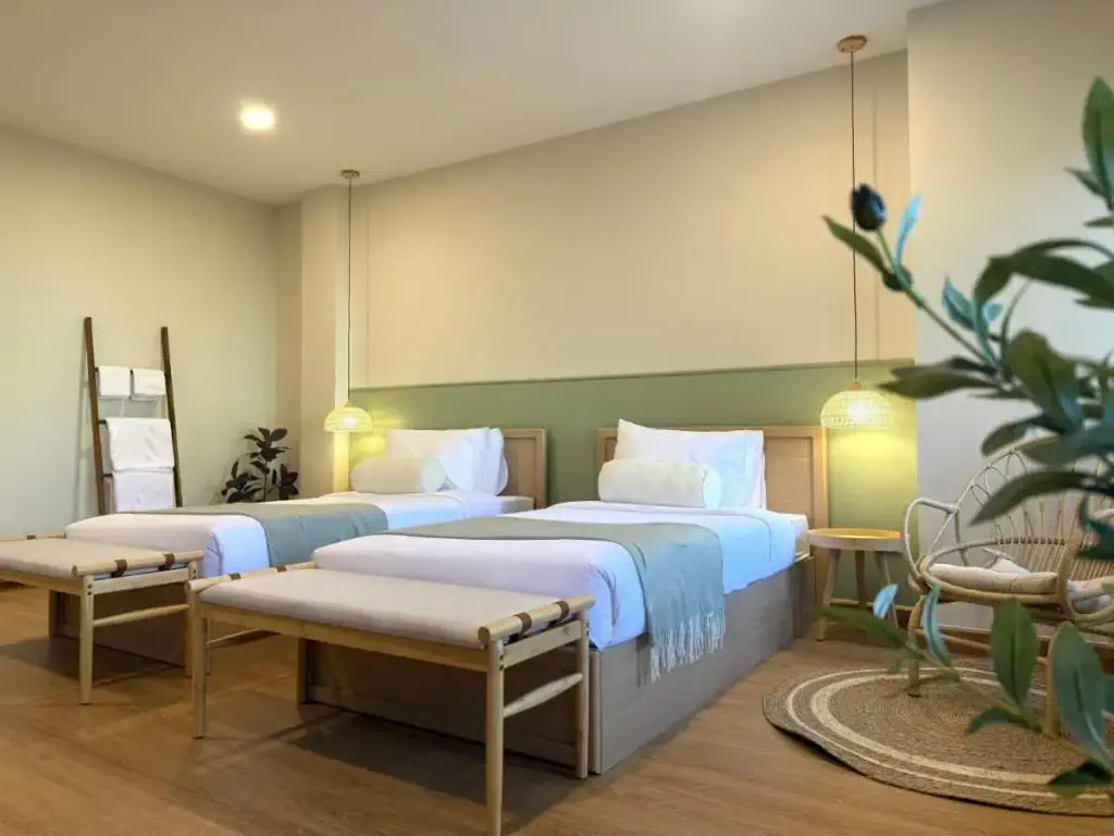 A cozy bedroom with two neatly made twin beds featuring light wood frames, white linens, and teal throws. Soft pendant lights hang above. A plant and wicker chair add a serene, natural touch.