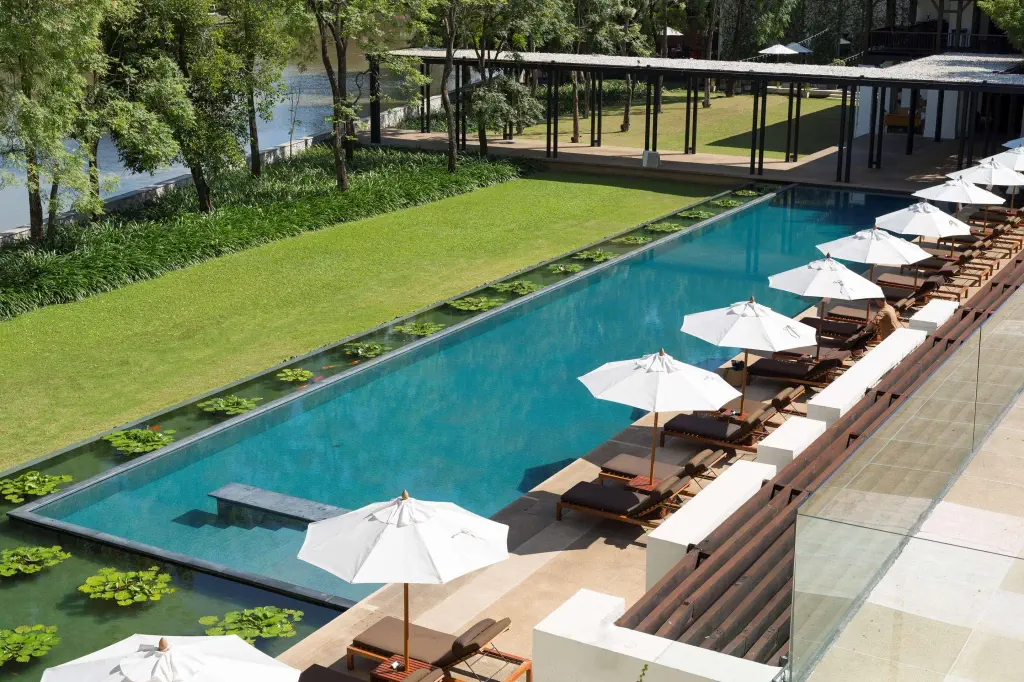 Luxurious outdoor swimming pool with clear blue water, bordered by sun loungers under white umbrellas. Surrounded by lush green lawn and trees.
