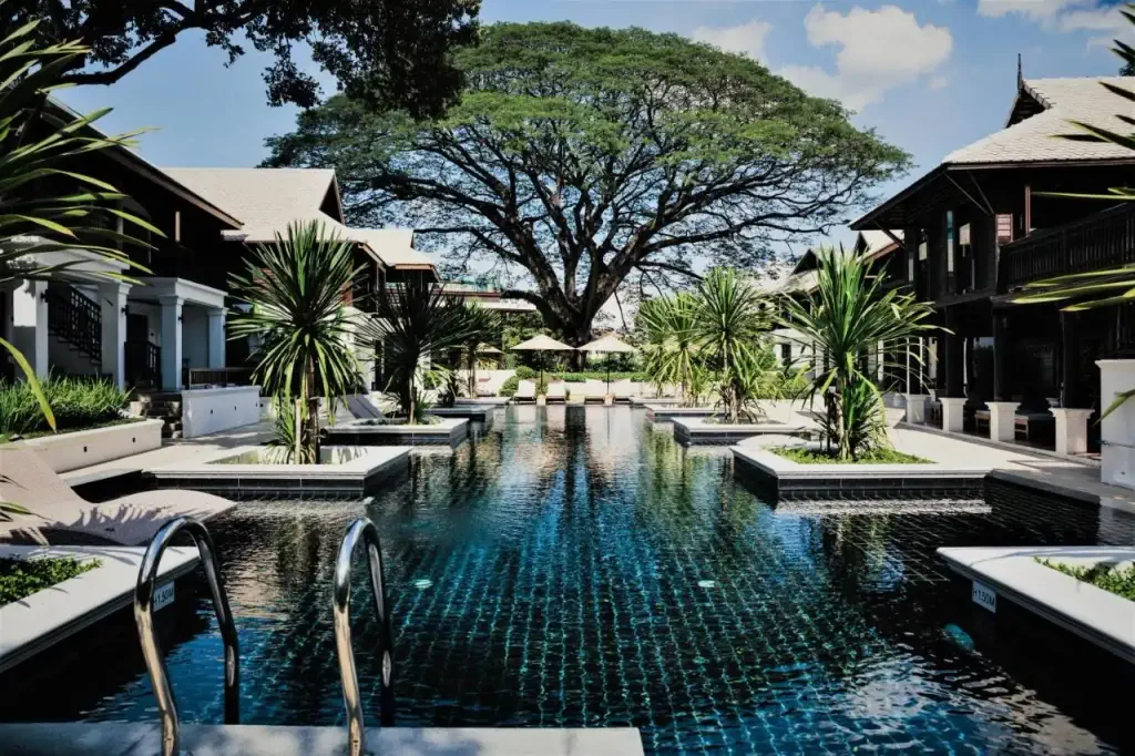Luxurious pool flanked by tropical plants and elegant buildings, leading to a majestic tree under a blue sky. The scene is serene and inviting.