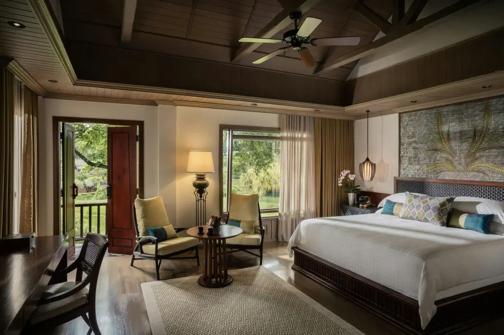 Luxurious bedroom with a large bed, patterned pillows, and a ceiling fan. A door and window open to lush greenery.