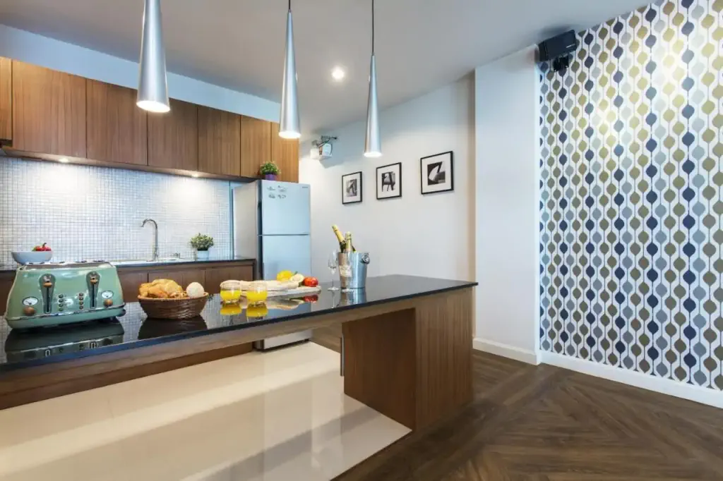 Modern kitchen with wooden cabinets, stylish pendant lights, patterned wallpaper, and a sleek island. Features a toaster, bread, fruit, and wine.