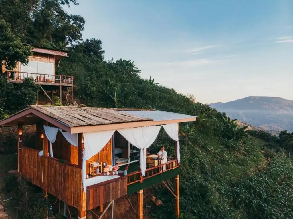 A wooden cabin with white curtains perched on a lush hillside, overlooking a vast mountain landscape under a clear blue sky. A person sits peacefully on the balcony.
