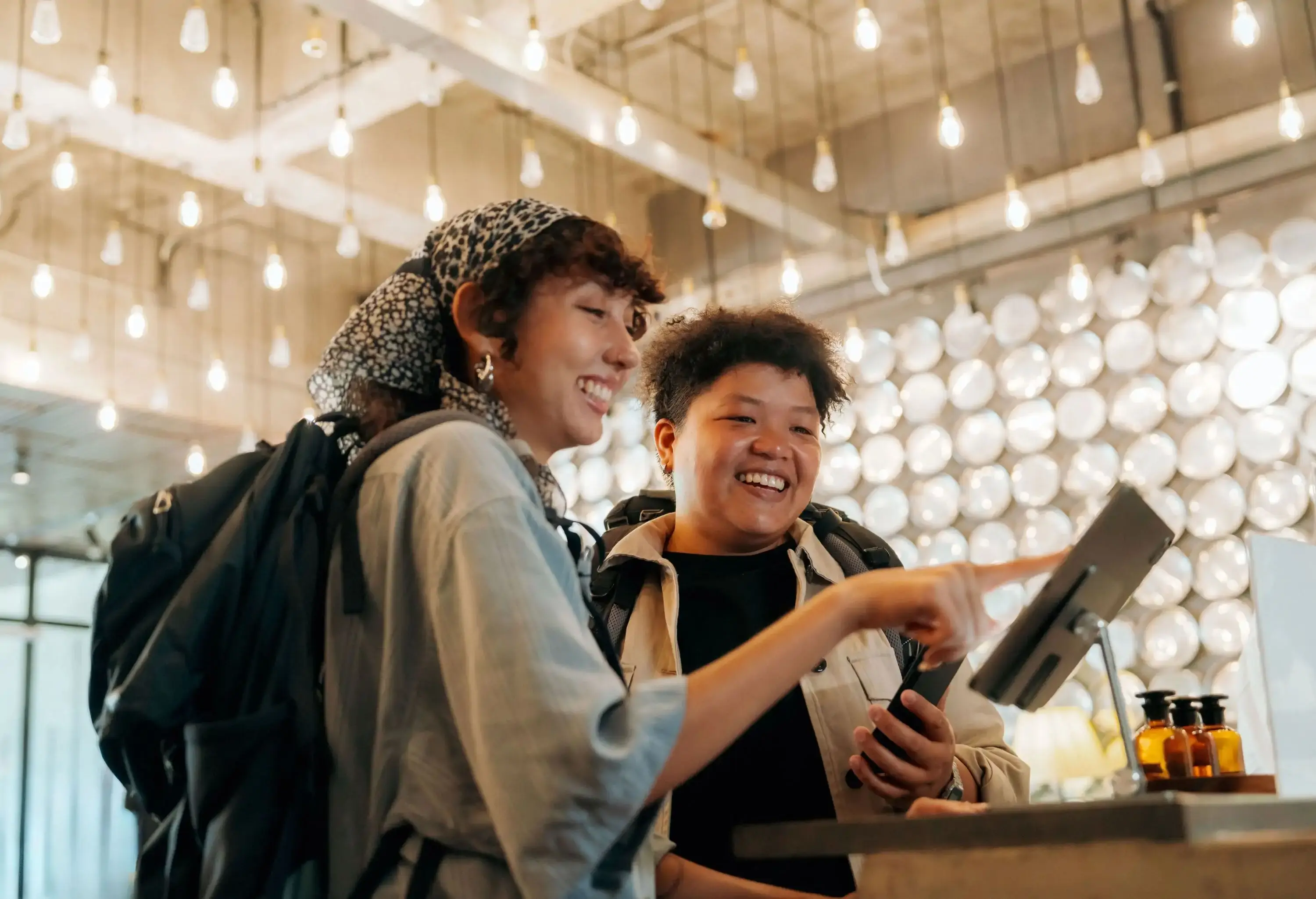 Two young adults smiling while using a self-service check-in system at a modern hostel. Concept of travel, friendship, and digital convenience in a trendy setting.