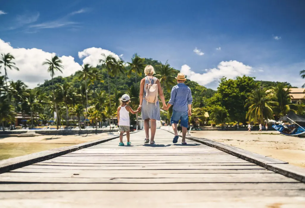 A woman and two young children holding hands as they walk toward the shore on a wooden boardwalk.