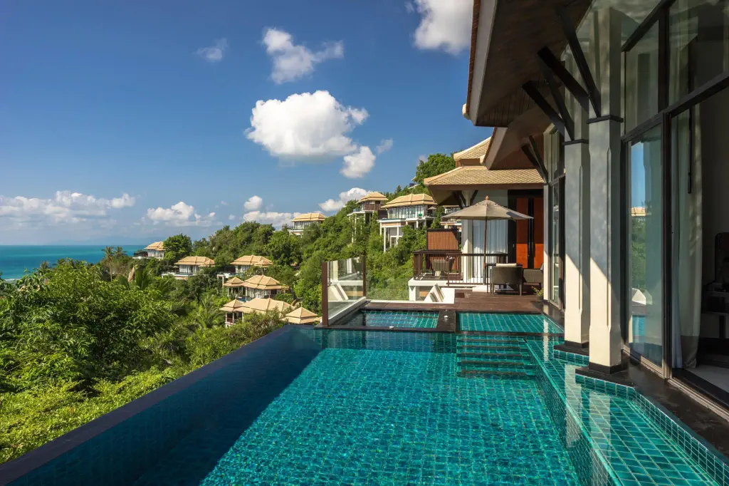 Luxurious hillside villa with infinity pool overlooking lush greenery and ocean. Bright, sunny day with blue sky and scattered clouds, creating a serene atmosphere.