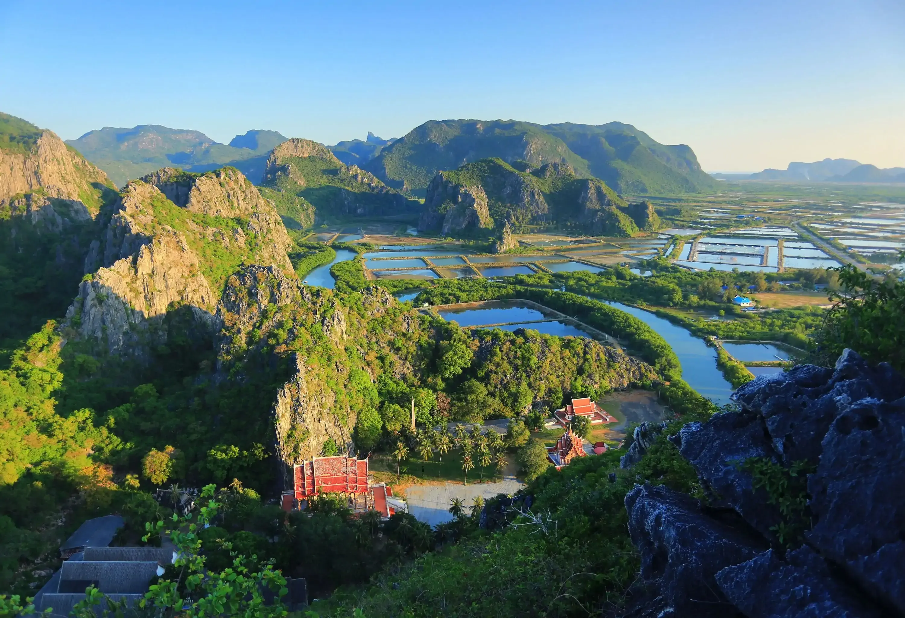 A temple in a mountainous landscape with jagged summits and lush trees along a wide field of rice paddies.