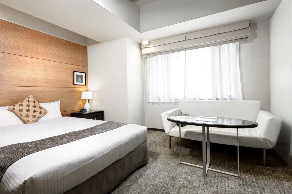 Modern hotel room with a double bed, bedside table with lamp, white sofa, glass coffee table, and large window with sheer curtains.