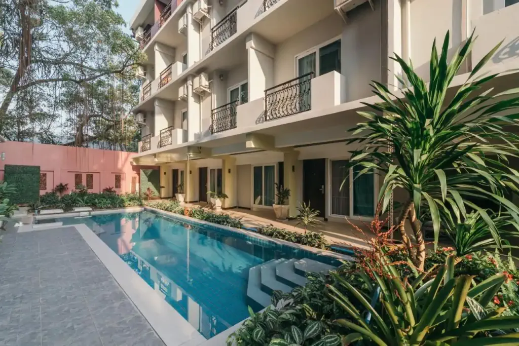 Modern hotel courtyard featuring a rectangular pool surrounded by lush plants. Multi-story building with balconies overlooks the tranquil setting.
