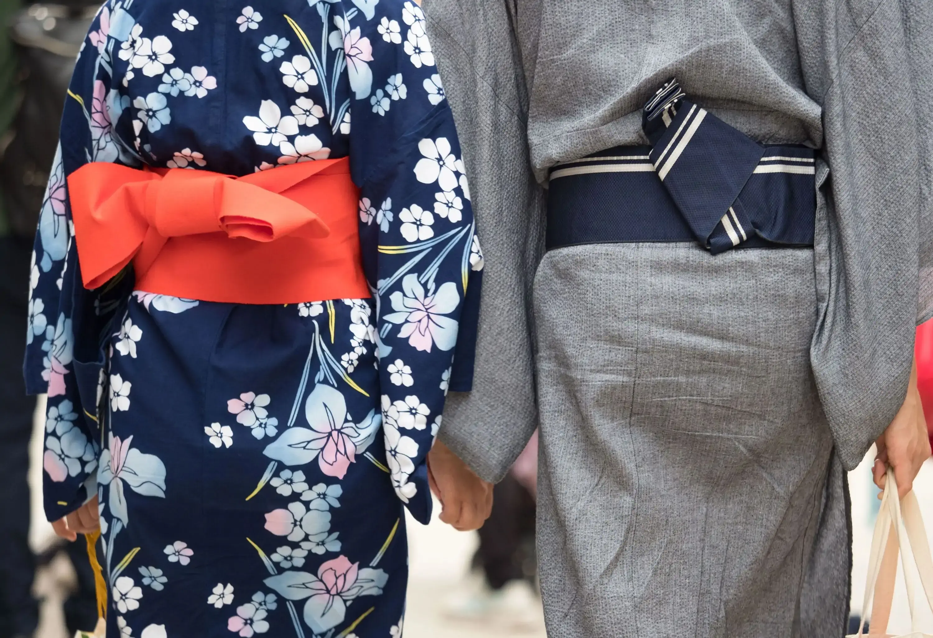 A couple wears floral kimono and grey yukata while holding hands.