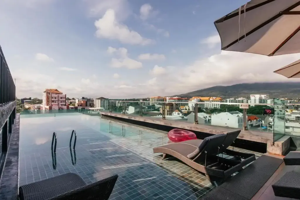 Rooftop infinity pool with a scenic cityscape and mountain view. Lounge chairs and umbrellas provide a relaxing atmosphere under a partly cloudy sky.