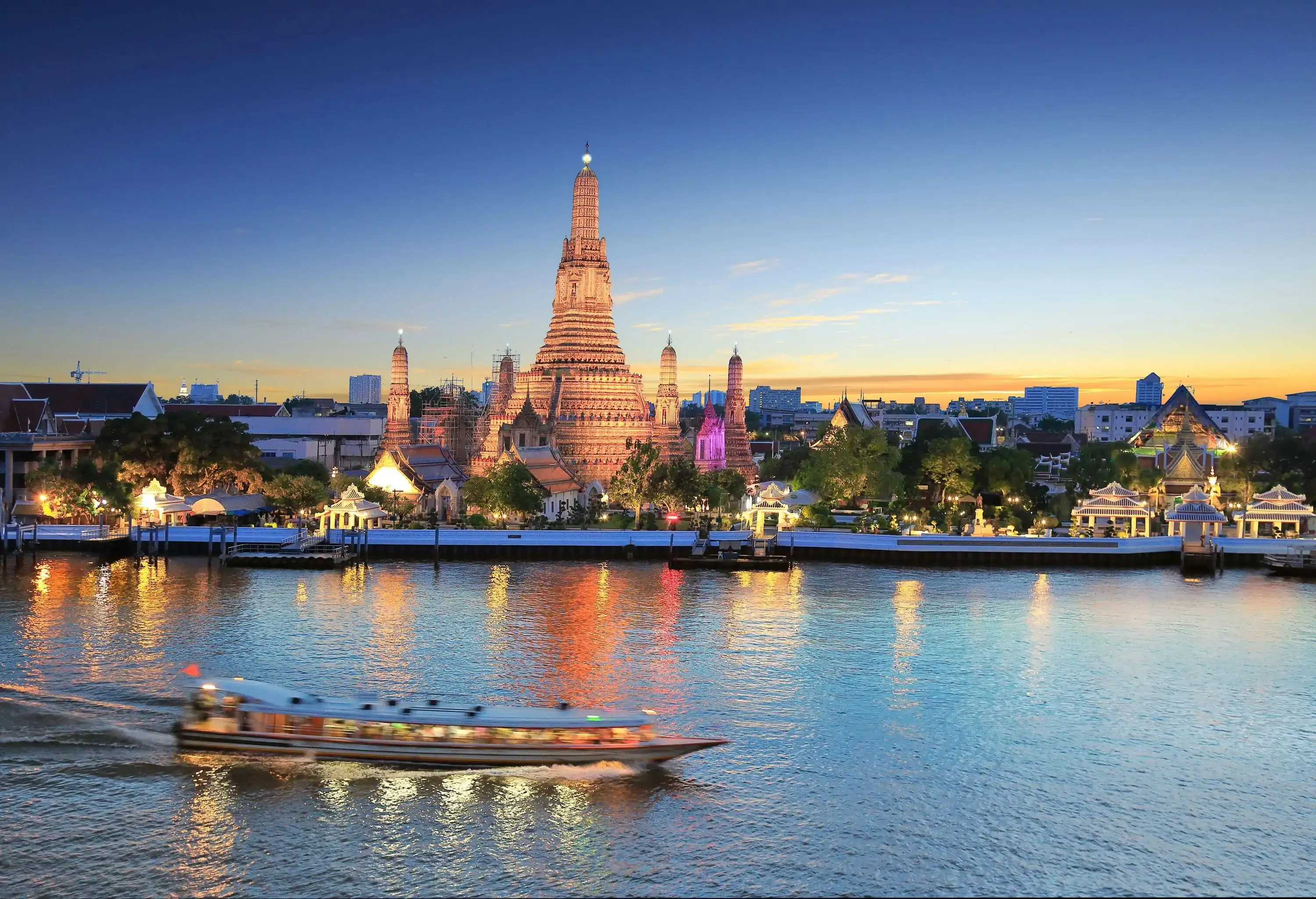 A passenger boat speeds through the river along the illuminated temples of Wat Arun during sunset.