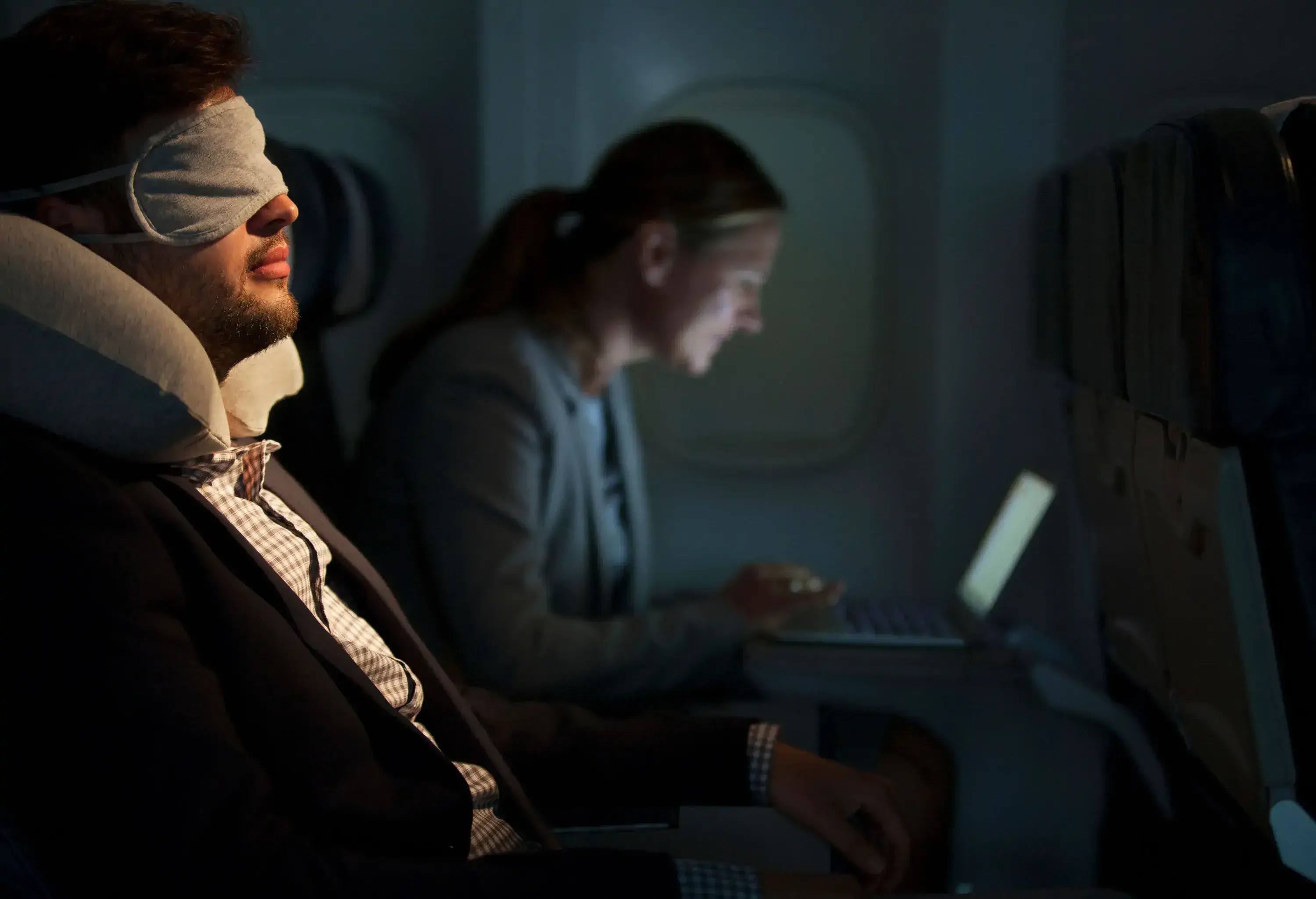 Man on a night flight sleeping with eye mask, whilst seat neighbour is on a mobile device