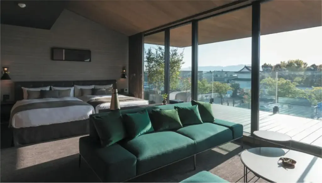 Luxurious bedroom with large windows, green sofa, and modern decor. Overlooks a sunny terrace with a scenic view.