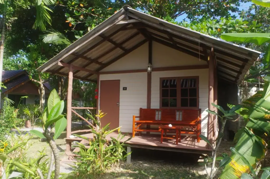 A small wooden cabin with a porch in a tropical setting. The cabin has a bench, surrounded by lush greenery and trees, conveying a peaceful vibe.