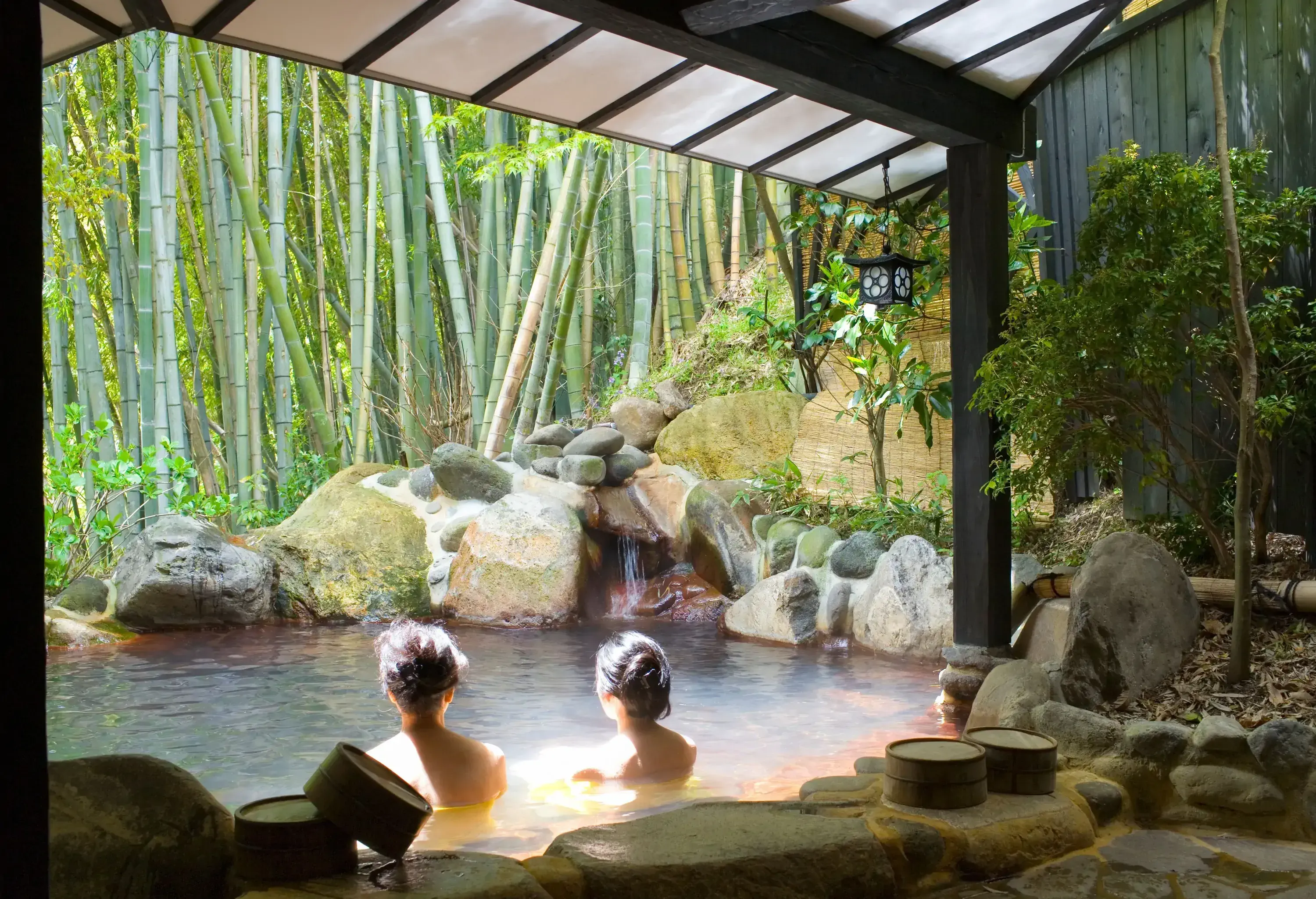 Two female friends submerged in an open-air hot-spring bath alongside the bamboo.