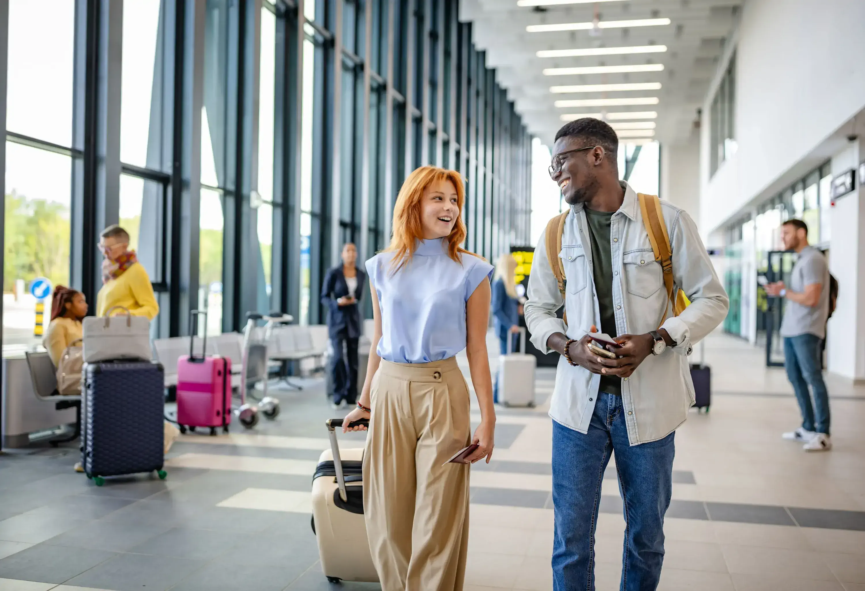 A man and woman walk confidently through an airport corridor, carrying rolling luggage, illustrating a vibrant travel moment filled with anticipation and conversation.