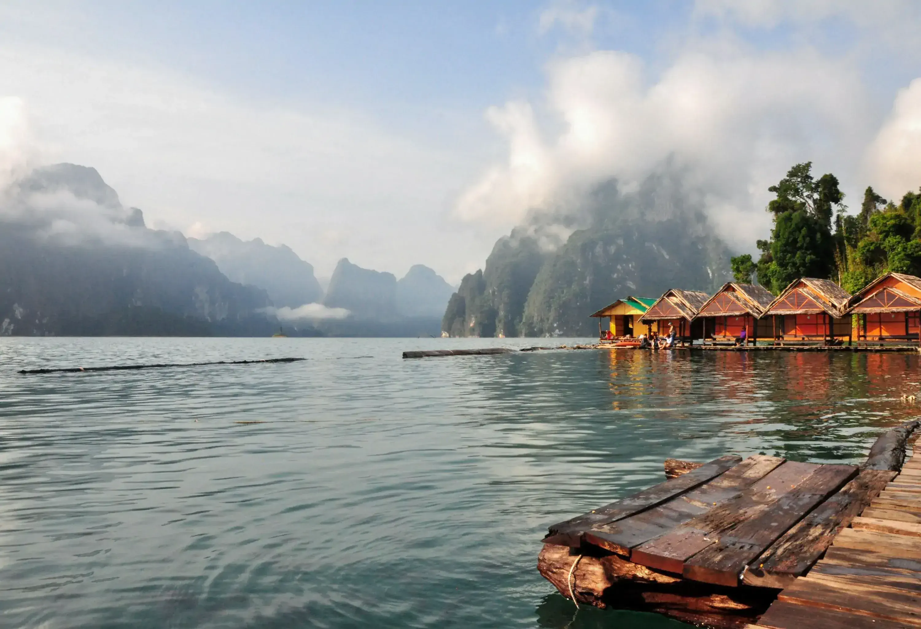 A tranquil lake lined with wooden houses and forested mountains surrounded by ​​clouds.