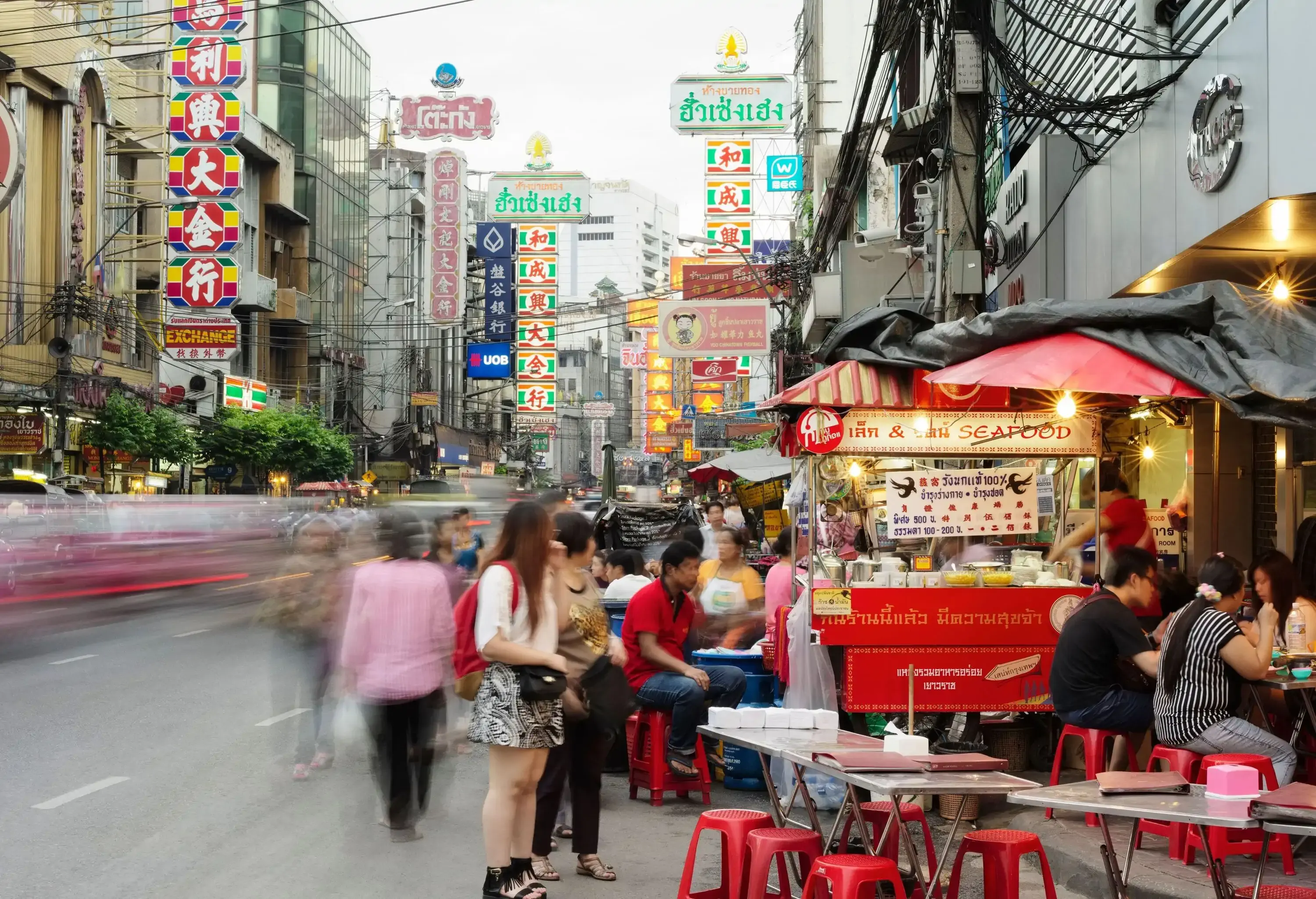 A crowded street with food stalls, seats, and tables packed with people walking and dining.