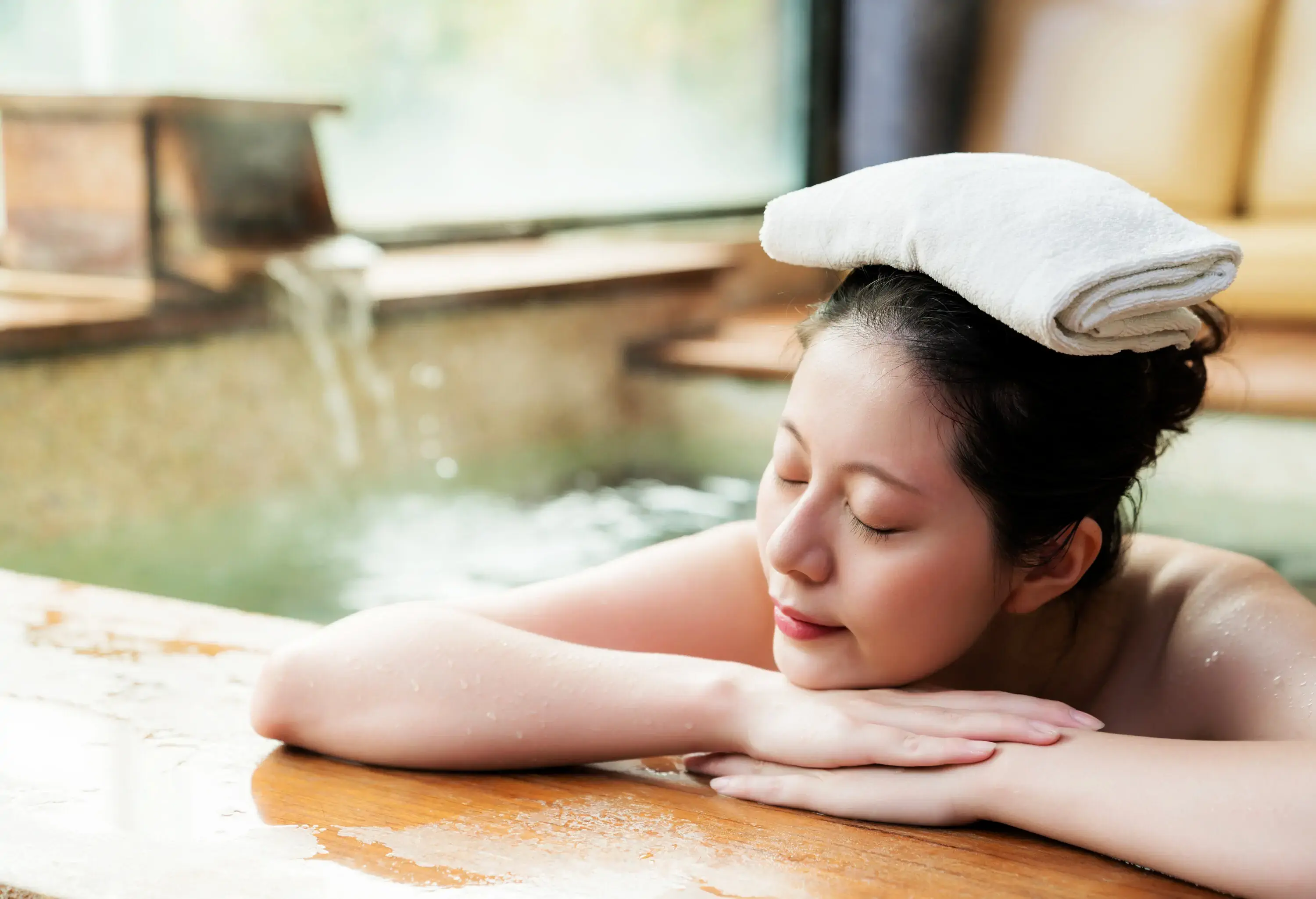A woman resting on the edge of a bath with her eyes closed and a folded towel over her head.