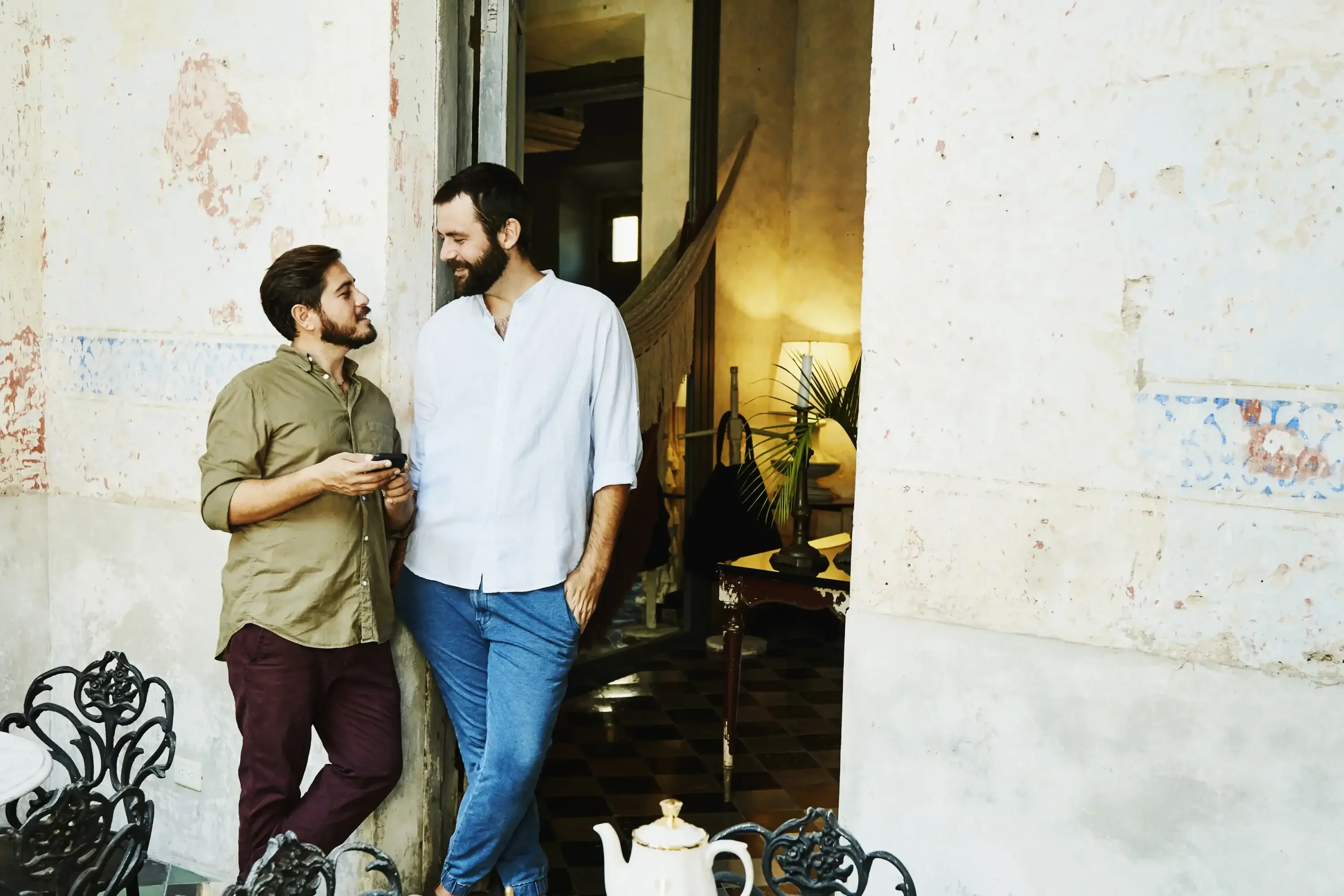 Two men stand in a rustic cafe, exchanging smiles. A wrought iron table with a teapot set is in the foreground. The atmosphere is relaxed and warm.