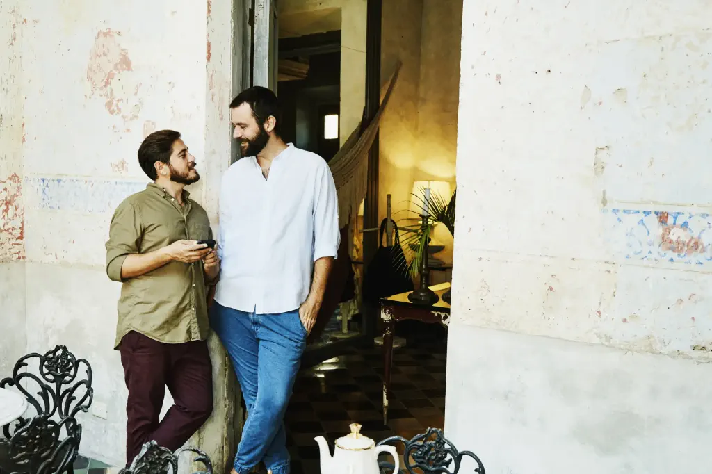 Two men stand in a rustic cafe, exchanging smiles. A wrought iron table with a teapot set is in the foreground. The atmosphere is relaxed and warm.