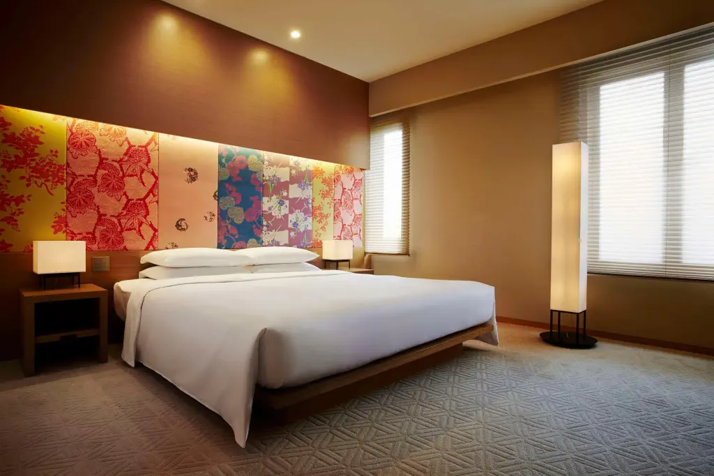Modern hotel room with a large bed facing an artistic floral panel. Soft lighting from windows and lamps creates a warm, inviting atmosphere.