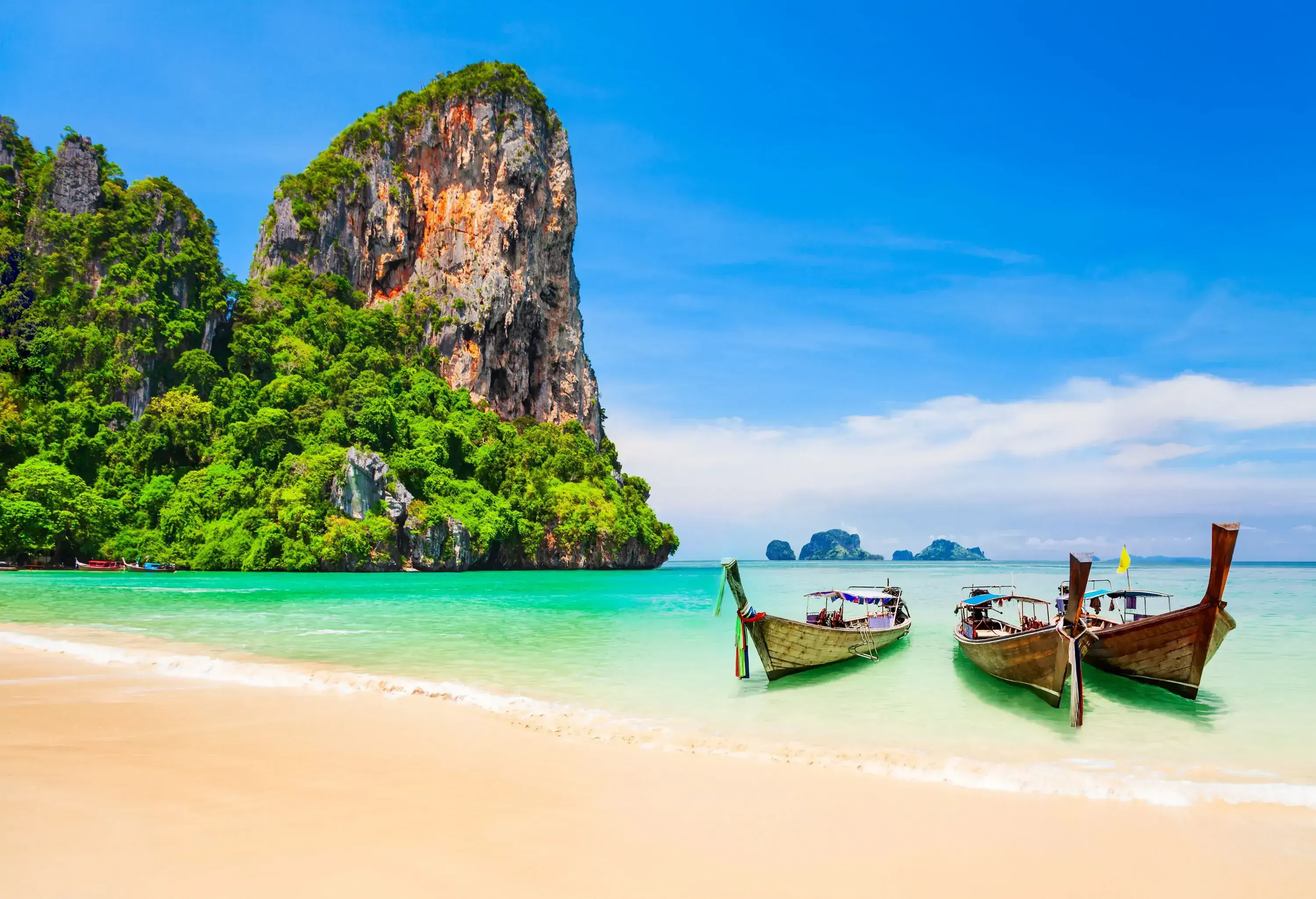 Three long-tail boats on the beach with steep-sided mountains in the backdrop.