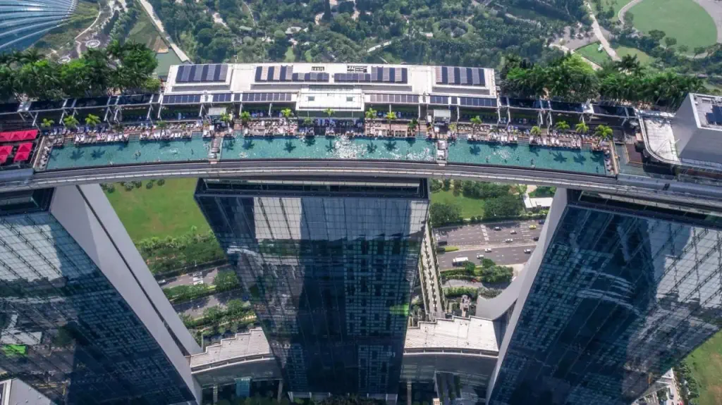 Aerial view of a large rooftop infinity pool atop three skyscrapers, surrounded by people and palm trees, with a cityscape and greenery in the background.